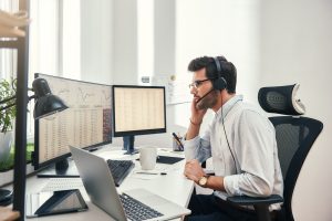 Side view of young bearded trader in headset talking with client and looking at monitor screen with trading charts and financial data while sitting in his modern office.