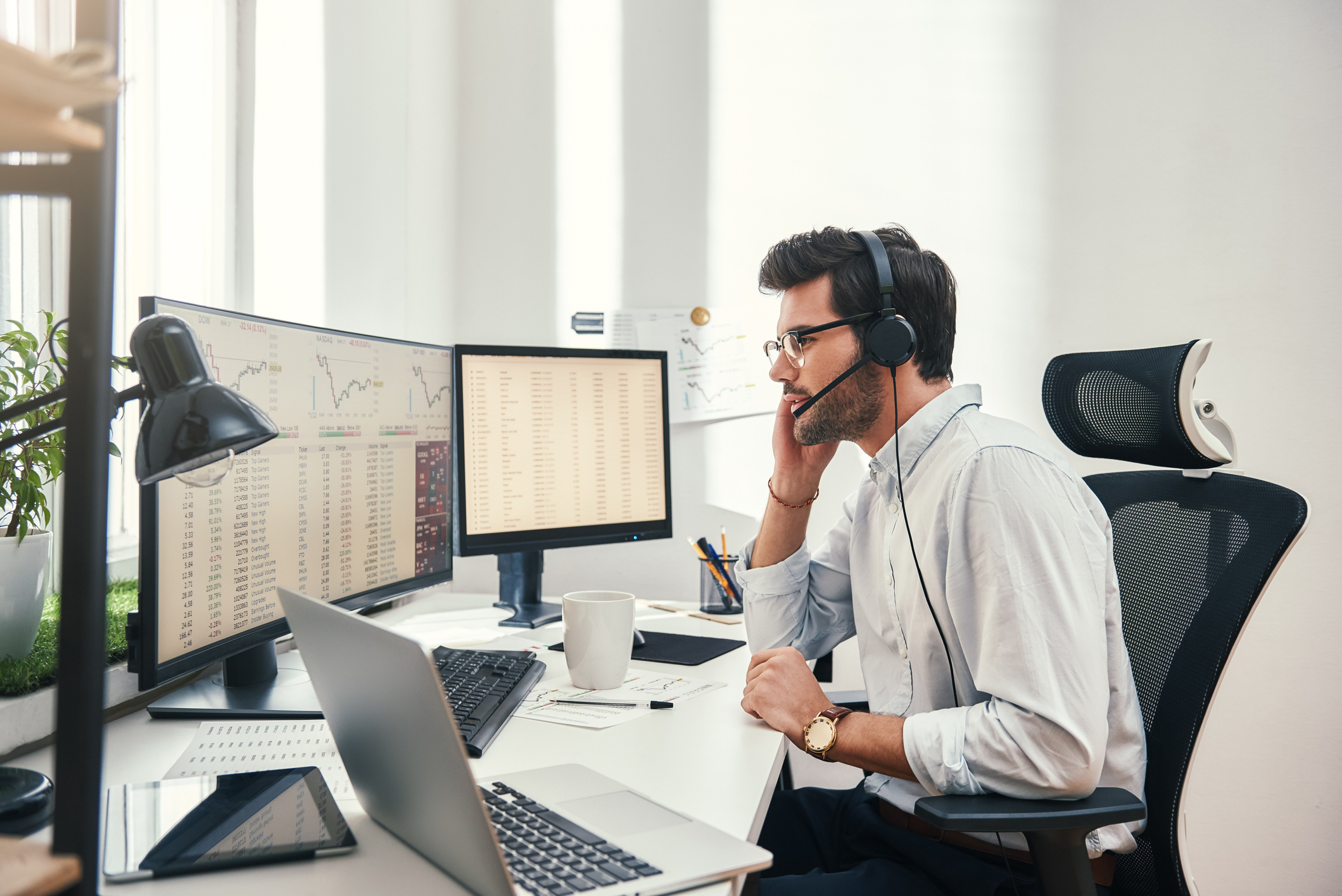 Side view of young bearded trader in headset talking with client and looking at monitor screen with trading charts and financial data while sitting in his modern office.